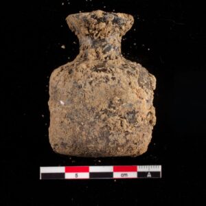 A small glass bottle, excavated at Jamestown, viewed from the side. The bottle is covered in dirt.