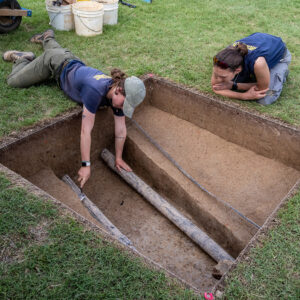 Staff Archaeologist Ren Willis scores the stratigraphic layers in one of the Smithfield excavation squares as Senior Staff Archaeologist Anna Shackelford looks on. The pipes traversing the square were installed in the 1930s and the network cables in the 2000s.