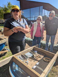 Archaeological Field Technician Hannah Barch shares artifacts found in the new excavation squares west of the Archaearium walking path. She is holding a partial Bartmann jug medallion.