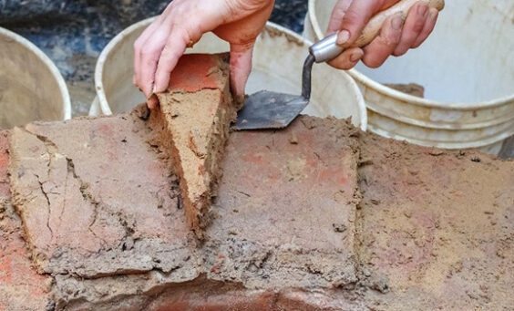 An archaeologist removes a wedge-shaped brick from the brick well ring using their hand and a trowel. The brick is covered in the original mortar.